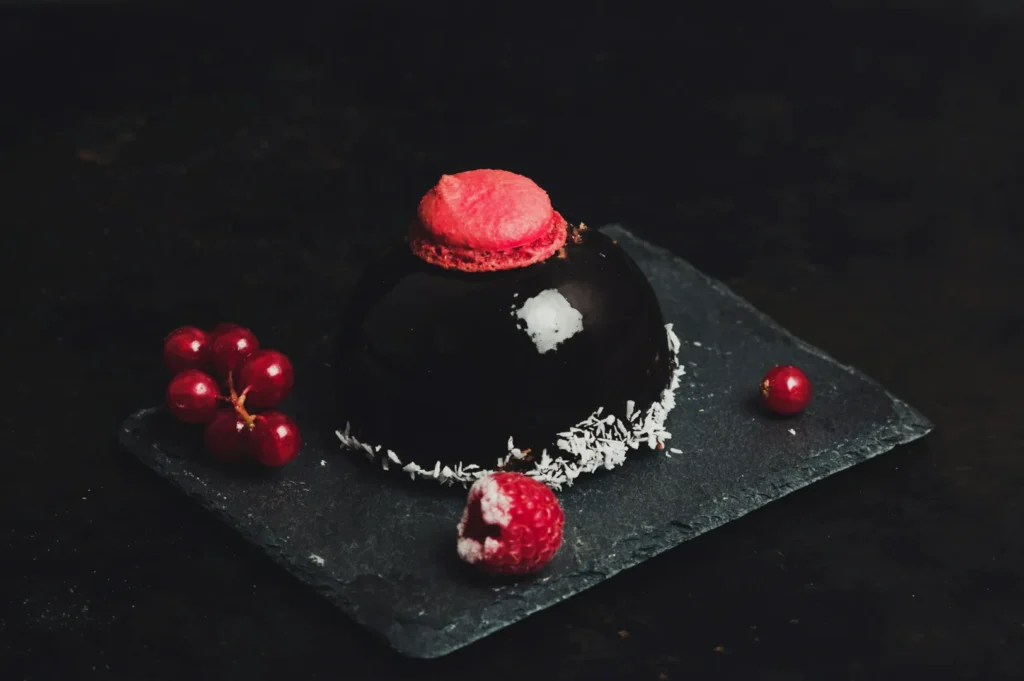 A glossy black chocolate dome dessert with coconut flakes, topped with a pink macaron, sits on a slate board. Surrounding are red currants and a sugared raspberry, creating an elegant and rich visual appeal.