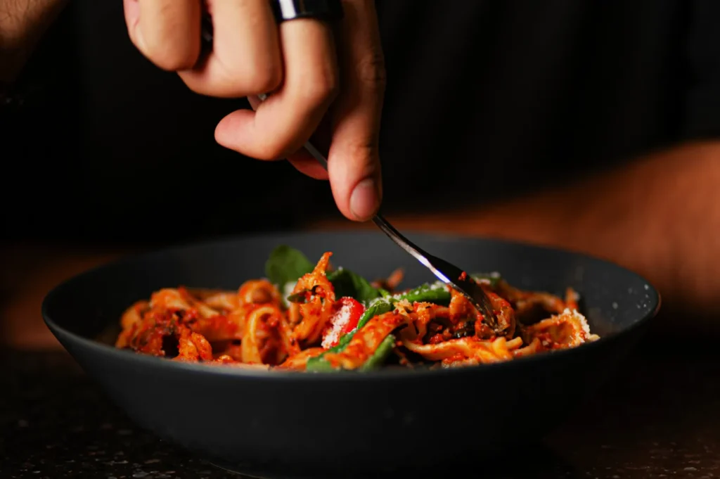 A hand twirls spaghetti with tomato sauce and greens in a dark bowl using a fork. The setting has a warm, inviting ambiance.