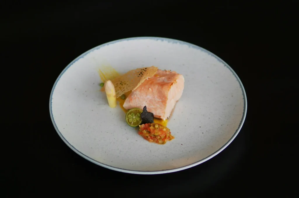 Elegant dish featuring a piece of cooked salmon topped with a lace garnish, asparagus, slices of lime, and a small scoop of salsa on a plain white plate.