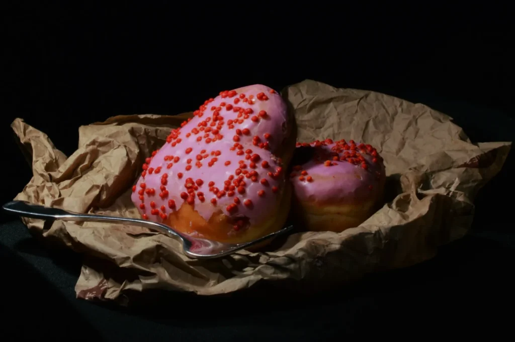 Pink-frosted heart-shaped donuts with red sprinkles rest on crumpled brown paper, accompanied by a fork, set against a dark background.