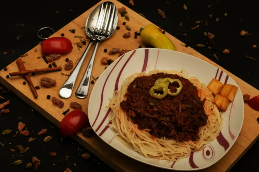 A plate of spaghetti topped with rich tomato sauce and sliced peppers, garnished with diced cheese. Set on a wooden board, with spices, tomatoes, and utensils. Warm, rustic ambiance.