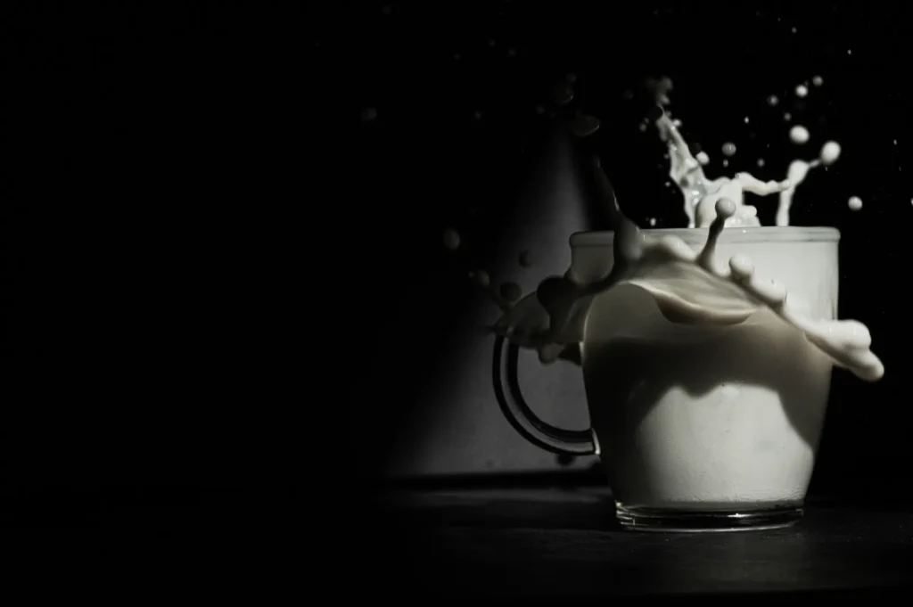 A white mug on a dark background with milk dramatically splashing out, creating arcs and droplets. The scene is dynamic, conveying motion and energy.