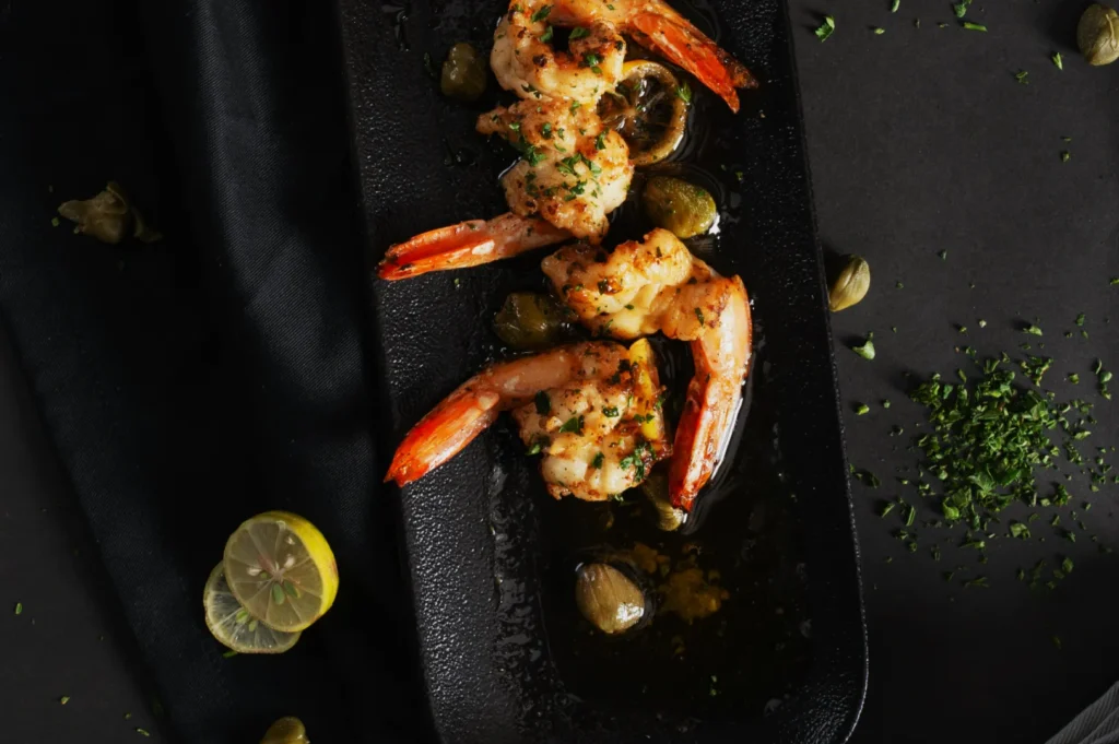 Grilled shrimp garnished with herbs on a black plate, surrounded by capers and lemon slices, creating an elegant, savory presentation.