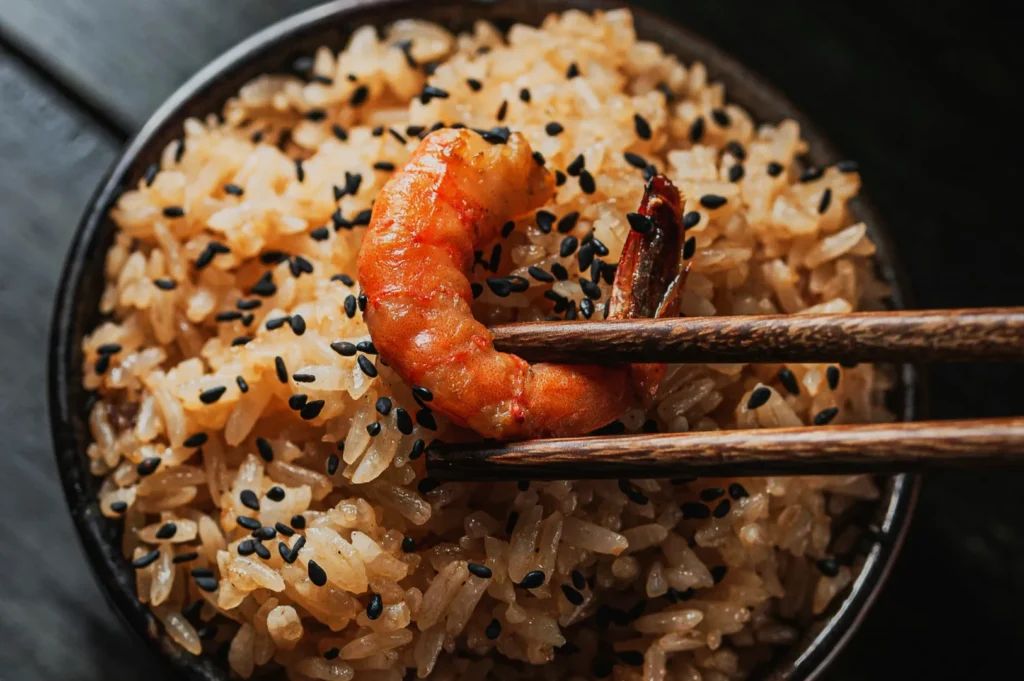 A close-up of a bowl of brown rice topped with black sesame seeds, with chopsticks holding a cooked shrimp. The dish looks savory and appetizing.