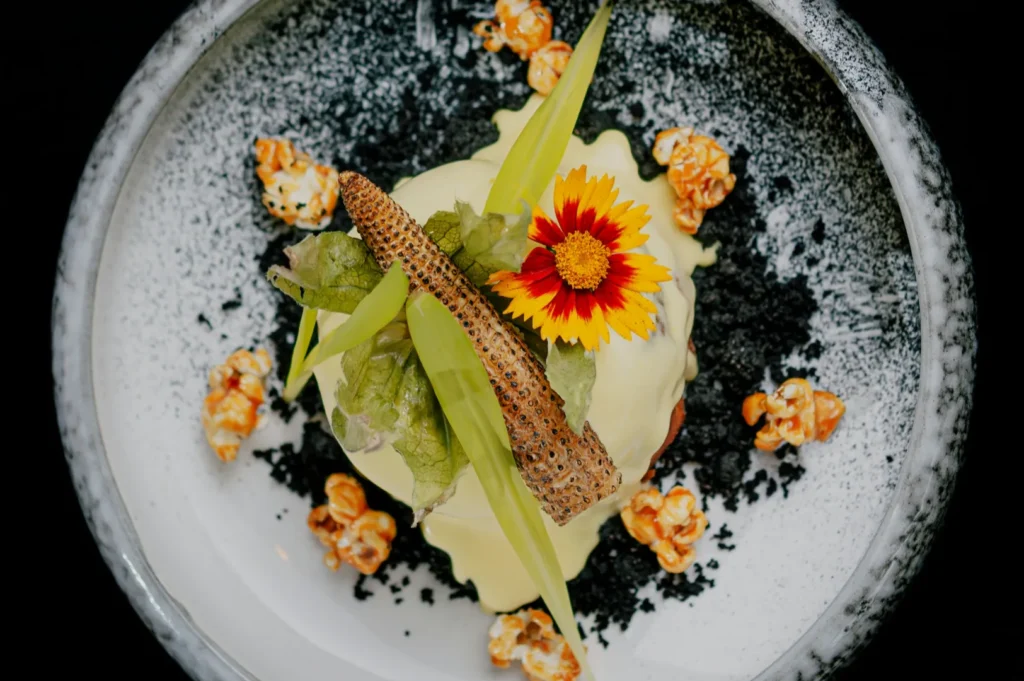 Gourmet dish with vibrant yellow sauce, a red and yellow flower, greens, crispy garnish on a speckled plate, surrounded by caramel popcorn.