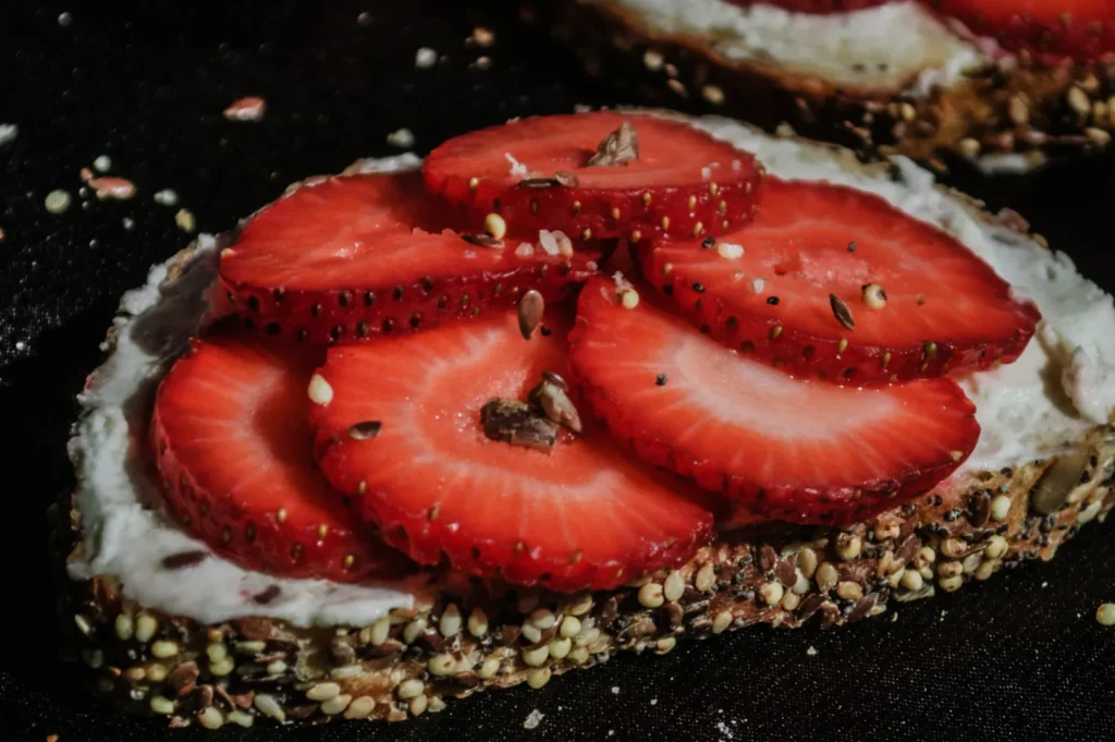 Close-up of multigrain bread topped with cream cheese and fresh strawberry slices, sprinkled with seeds, on a dark surface, creating a rustic, appetizing look.