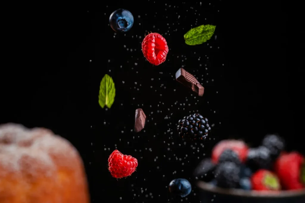A pink macaron balanced on a smartphone, with another underneath on a stand. Powdered sugar sprinkles create a whimsical, artistic scene on a black background.
