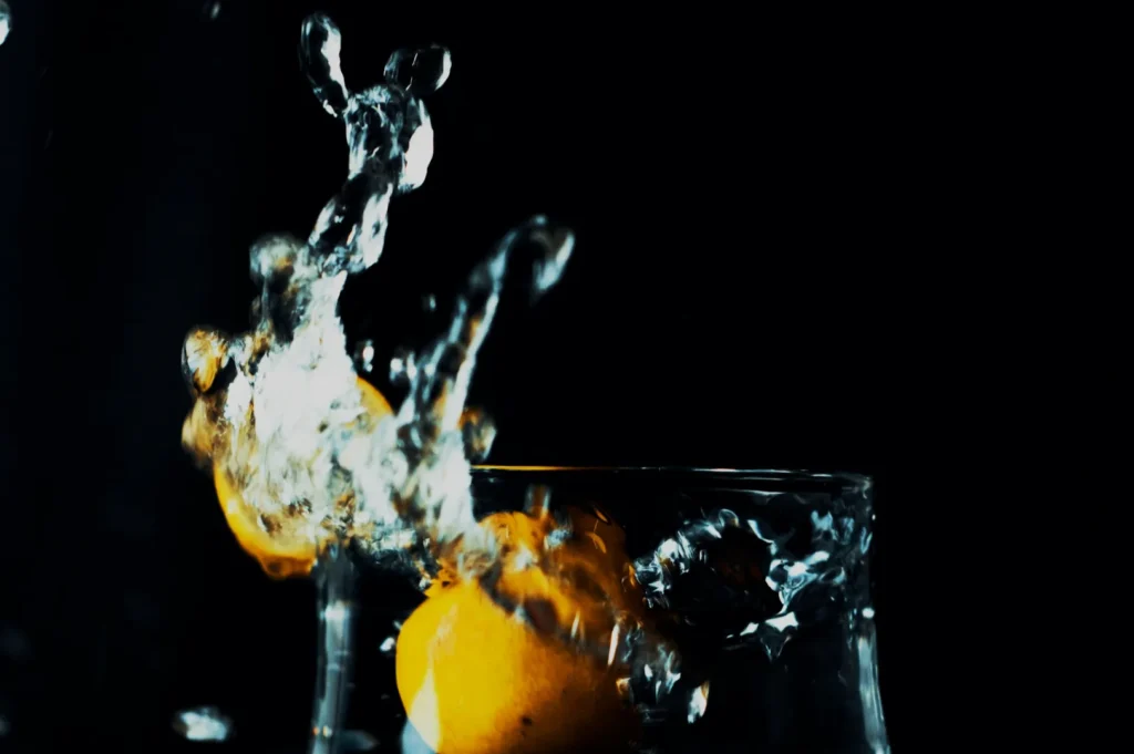 A lemon splashes into a glass of water, sending droplets and ripples upward. The image captures the motion mid-splash against a dark background.