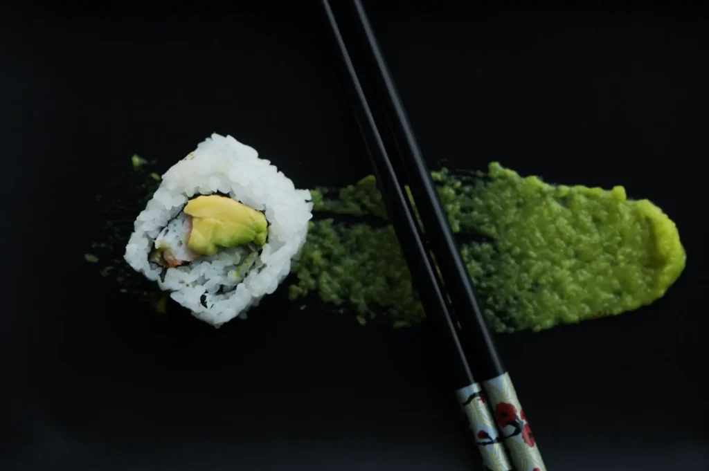 A sushi roll with avocado on a black plate, accompanied by a smear of green wasabi. Black chopsticks cross over the wasabi, creating a minimalist design.