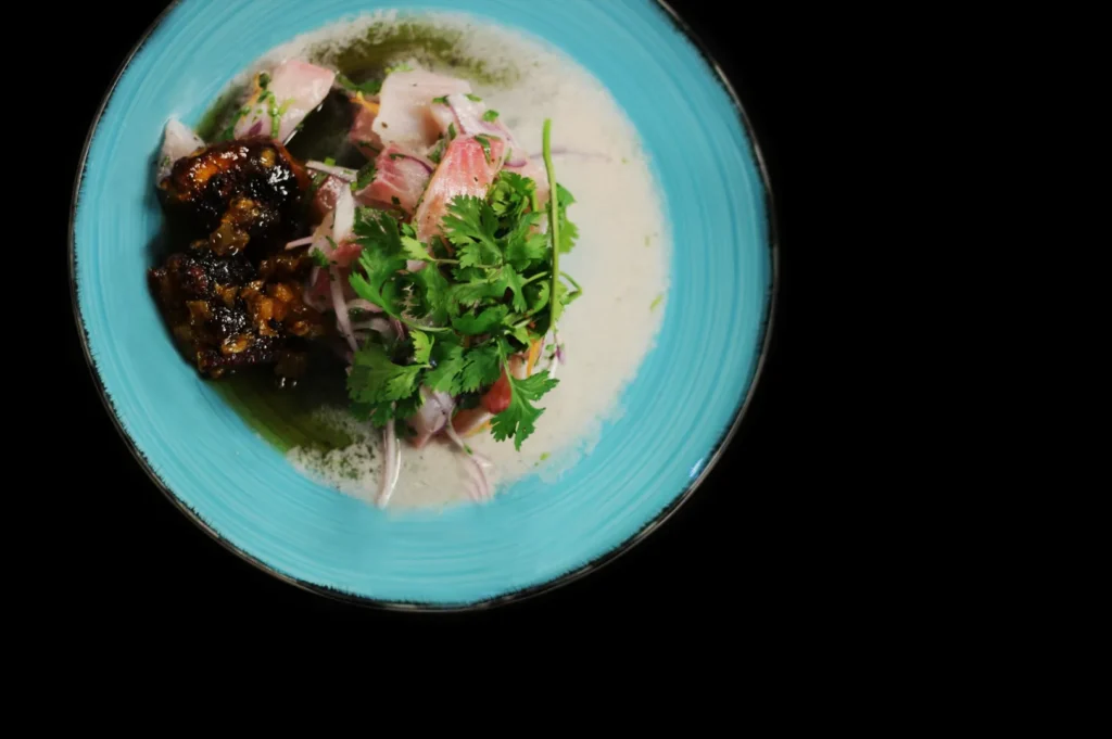 A vibrant dish on a turquoise plate features fresh ceviche with onion slices, cilantro garnish, and crispy pork. The presentation is elegant and enticing.