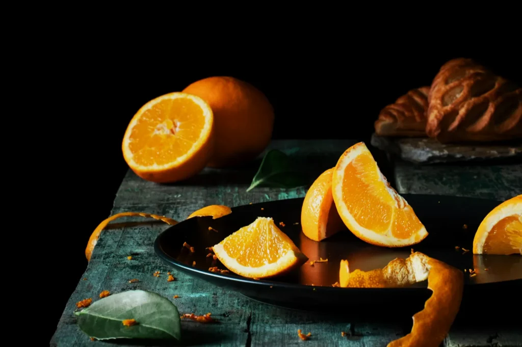 Slices of vibrant orange with peel on a black plate, set on a rustic blue surface. In the background, a croissant on a wooden board. Minimalist and fresh.