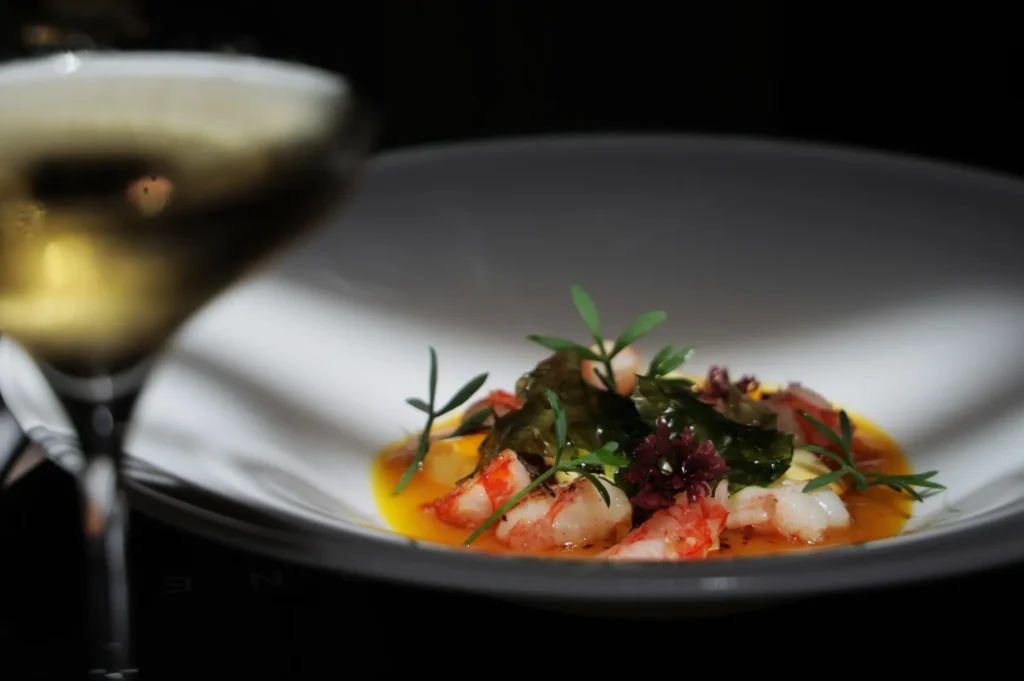 Gourmet dish with prawns, vibrant herbs, and greens in a glossy sauce on a white plate. A blurred glass of white wine is in the foreground.