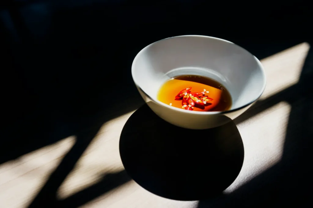 A white bowl holds brown sauce with sliced red chilies, bathed in dramatic light and shadow on a wooden surface, evoking a modern, minimalist feel.