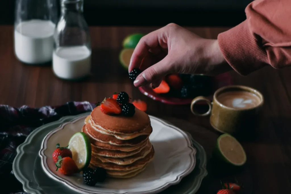 A hand places a blackberry on a stack of pancakes topped with strawberries and lime on a plate. Nearby are milk bottles and a cup of coffee.