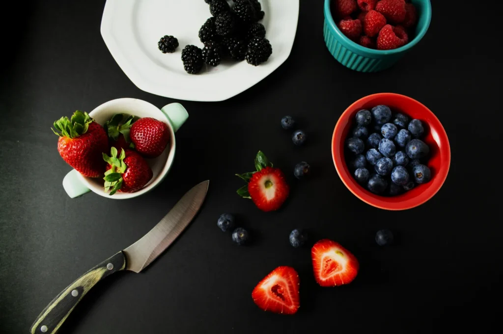 Assorted berries on black background: strawberries in a cup, blackberries on a plate, raspberries in a bowl, and blueberries in a red bowl with a knife nearby.