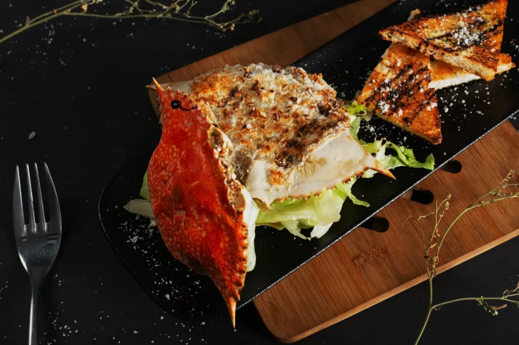 Grilled crab served on a black platter with crispy toast slices, garnished with lettuce. The setting is rustic with a dark, elegant ambiance.