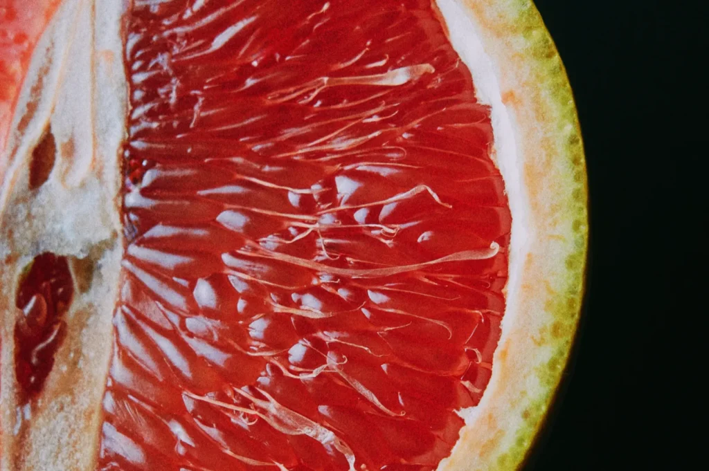 Close-up of a ripe grapefruit half against a dark background. The juicy, red pulp glistens, contrasting with its pale yellow rind. Fresh and vibrant.