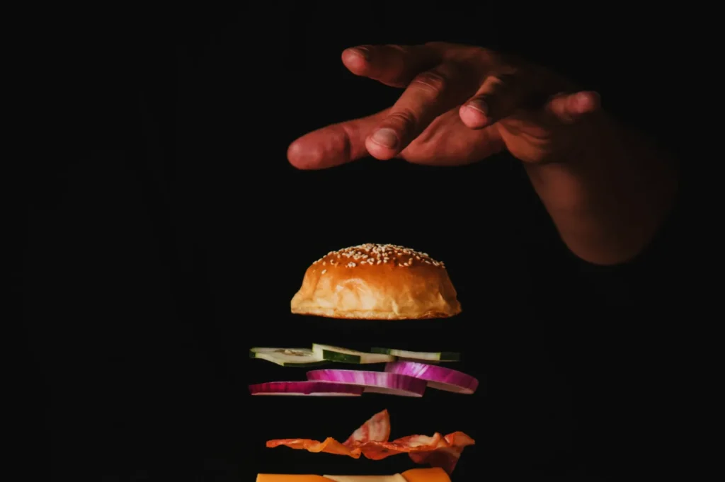 Floating burger layers in mid-air with a sesame bun on top, cucumbers, onions, bacon, and cheese beneath. A hand hovers above, dark background.