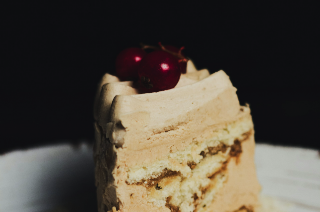 A slice of layered cake with beige frosting on a white plate, topped with two red berries. The dark background creates a rich, indulgent feel.