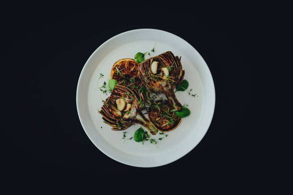A white plate features three grilled artichoke halves topped with garlic cloves and fresh herbs. The minimalist presentation is elegant and appetizing.