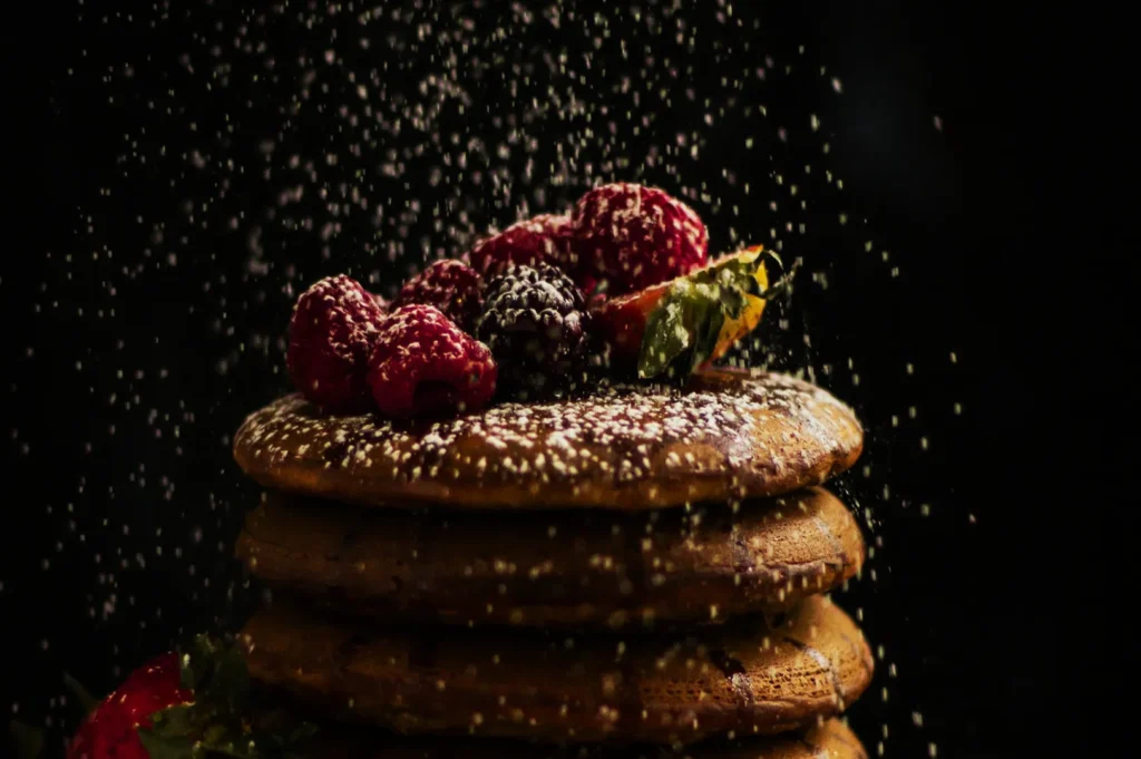 Stack of pancakes topped with raspberries and blackberries, dusted with powdered sugar on a dark background, creating a cozy, indulgent mood.