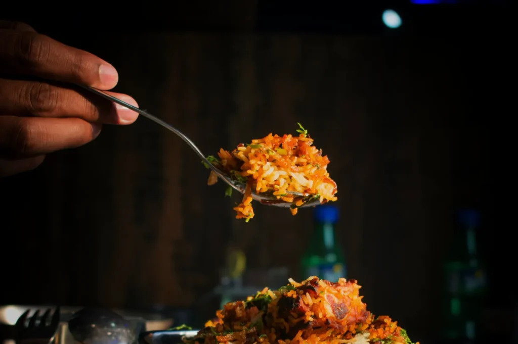 A close-up of a hand holding a spoonful of colorful biryani, garnished with herbs, above a plate. Dimly lit setting with a cozy, inviting mood.
