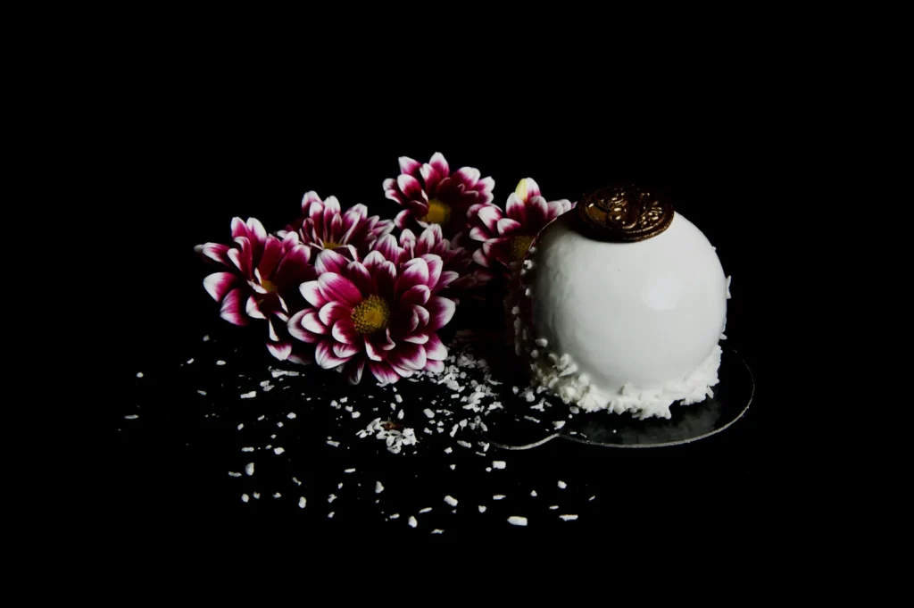 A white, glossy dessert topped with a chocolate disk is surrounded by pink and white flowers on a dark background. Coconut shavings add texture.