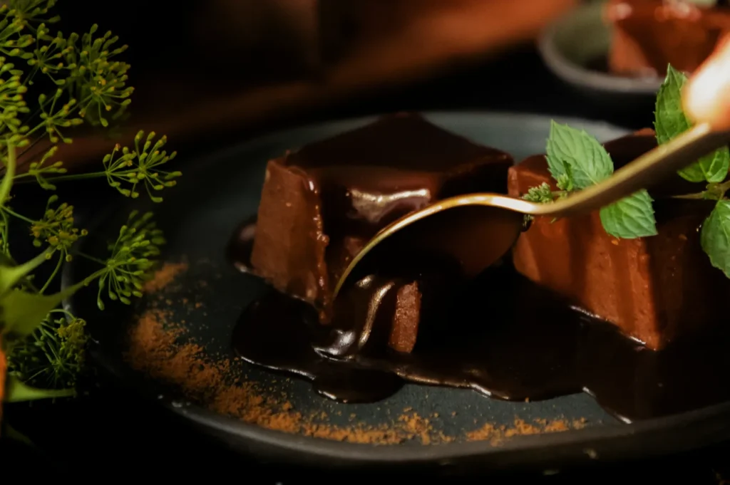 Chocolate dessert covered in glossy sauce with a golden spoon on a dark plate, garnished with fresh mint leaves and surrounded by greenery. Moody and indulgent.