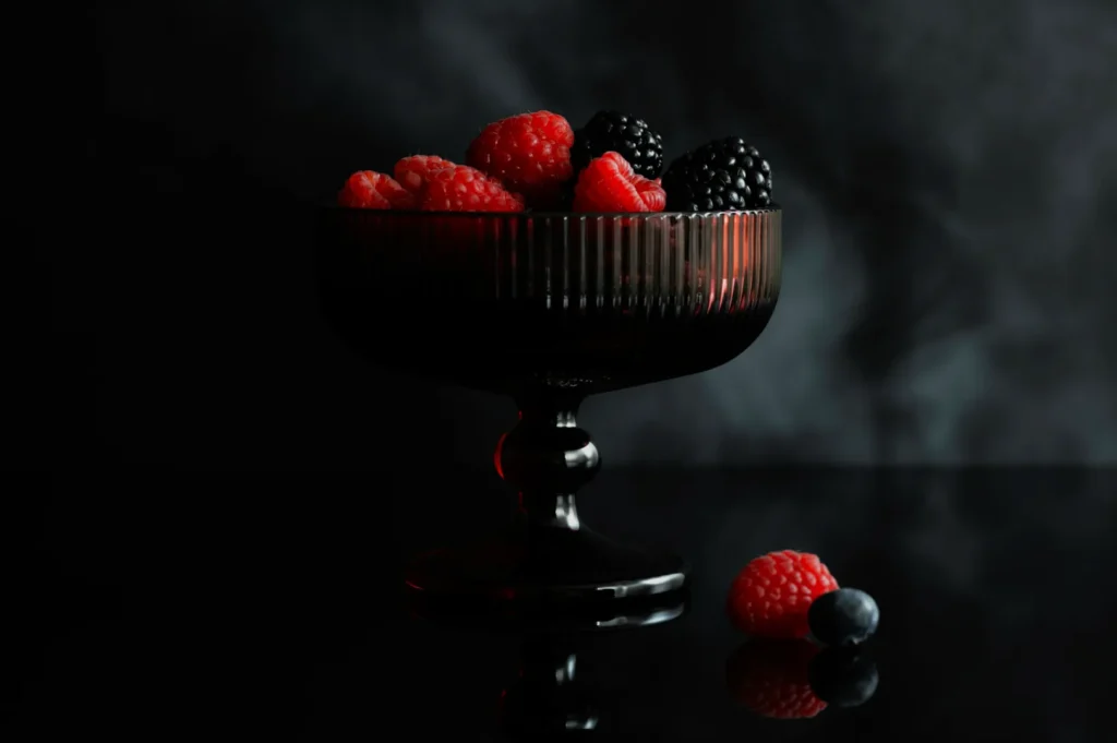 A dark glass goblet filled with raspberries and blackberries against a smoky black background. A few berries are scattered beside the goblet on a reflective surface, conveying a refined and moody tone.