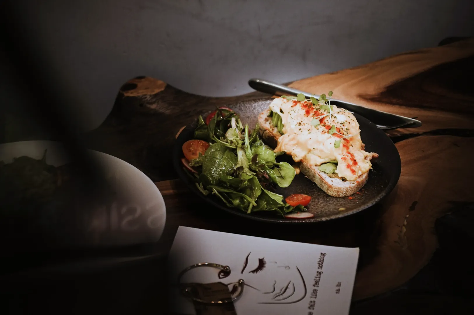 A plate with an open-faced sandwich topped with scrambled eggs and herbs sits beside a fresh green salad with cherry tomatoes on a rustic table. The setting feels cozy and artistic, with a sketch of a face partially visible on a paper next to the plate.
