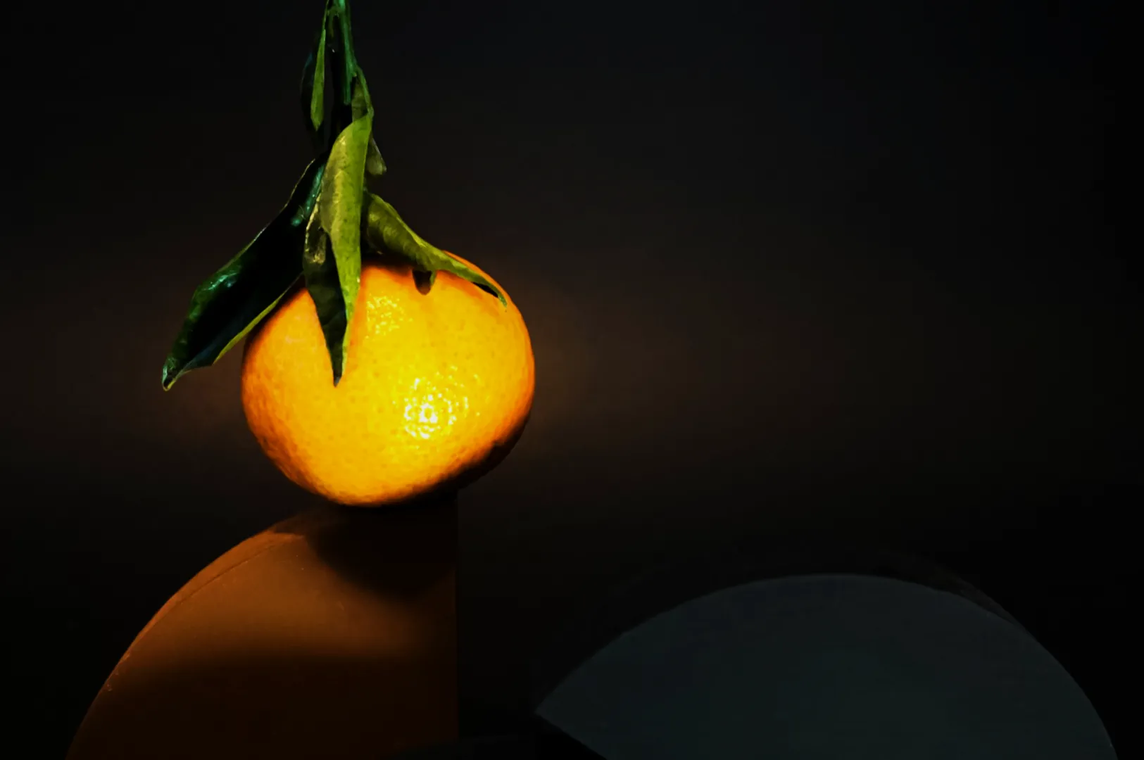 A ripe orange with glossy leaves rests on an angular, geometric surface against a dark background, creating a minimalist and elegant composition.