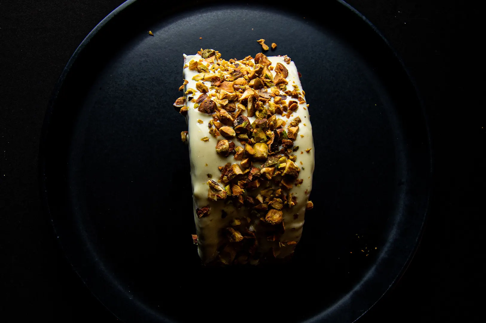A delicious dessert topped with white cream frosting and chopped pistachios served on a black plate.