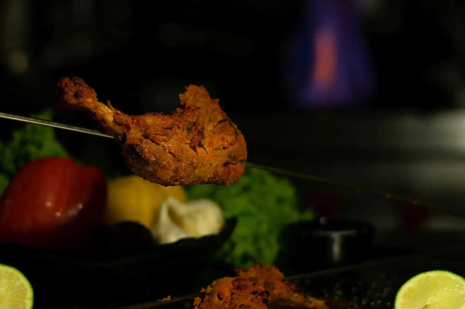Grilled tandoori chicken leg piece on a skewer with fresh lime and vegetables.