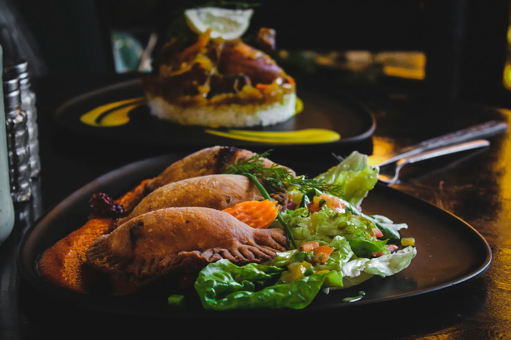Three golden-brown empanadas served on a black plate with a fresh side salad and dipping sauce.