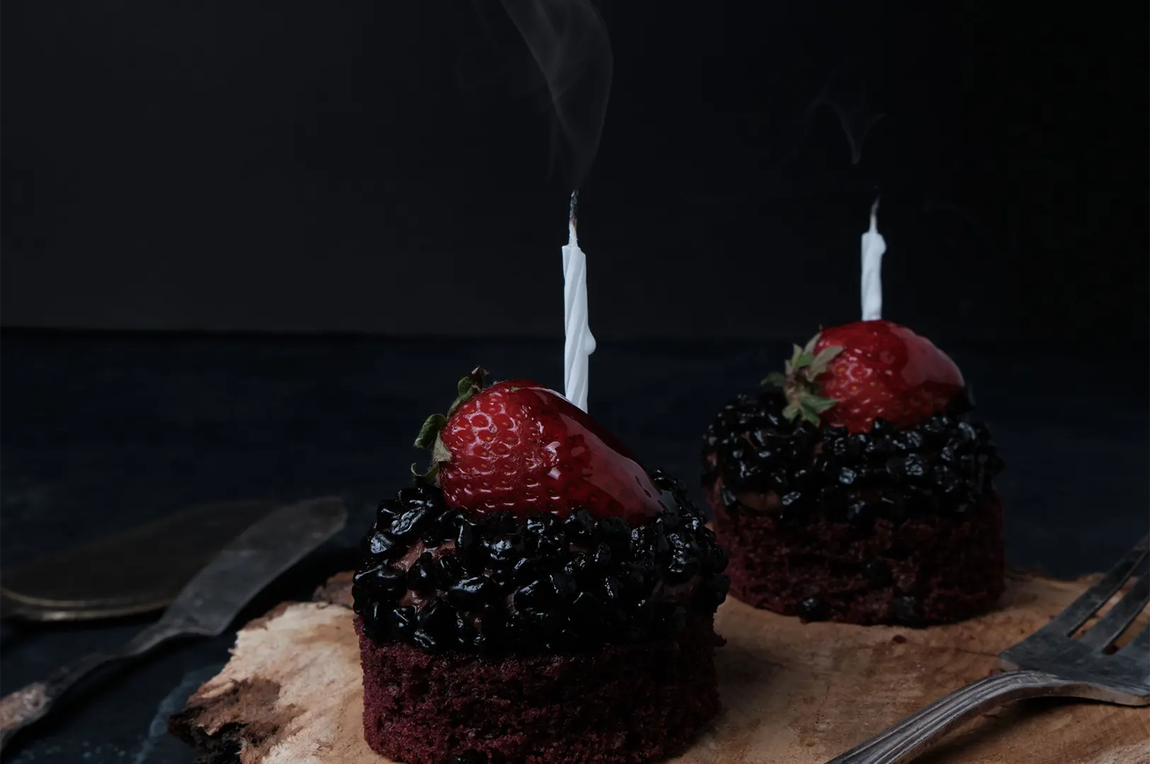 Food photography of two mini chocolate birthday cakes with strawberries, sprinkles, and lit candles on a wooden board.