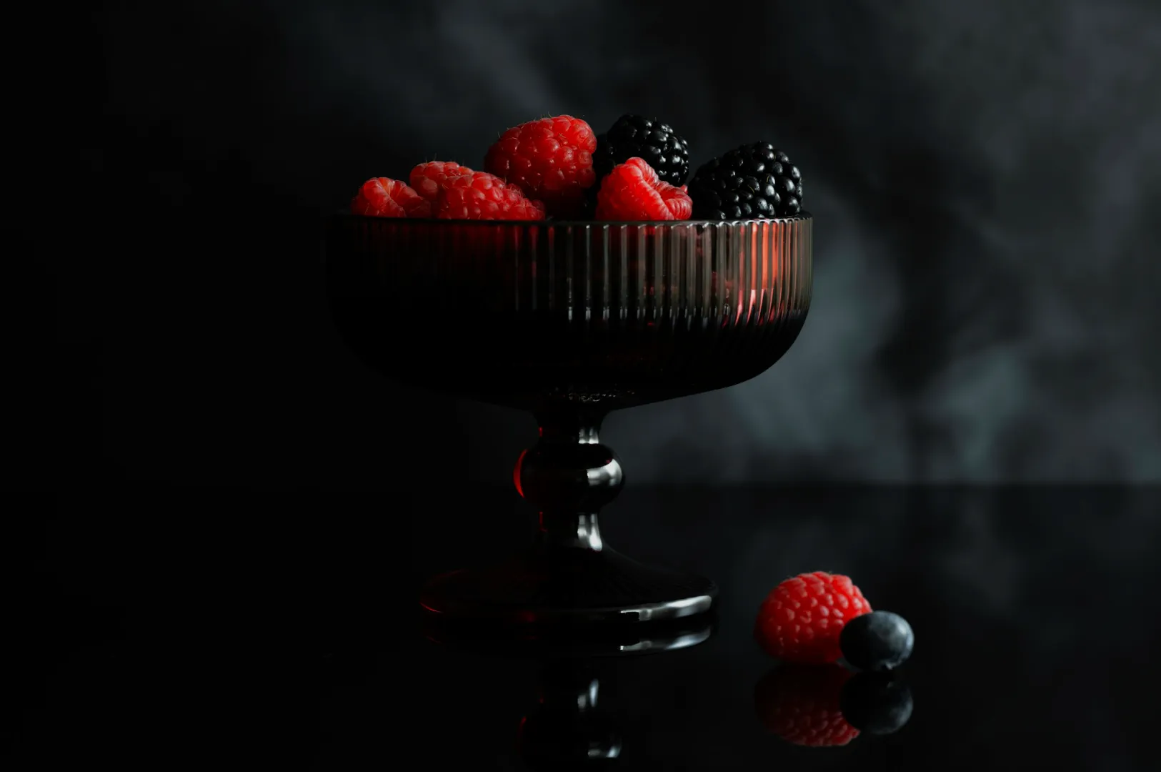 A dark glass bowl holds vibrant raspberries and blackberries against a smoky background. A raspberry and blueberry lie beside, creating an elegant contrast.