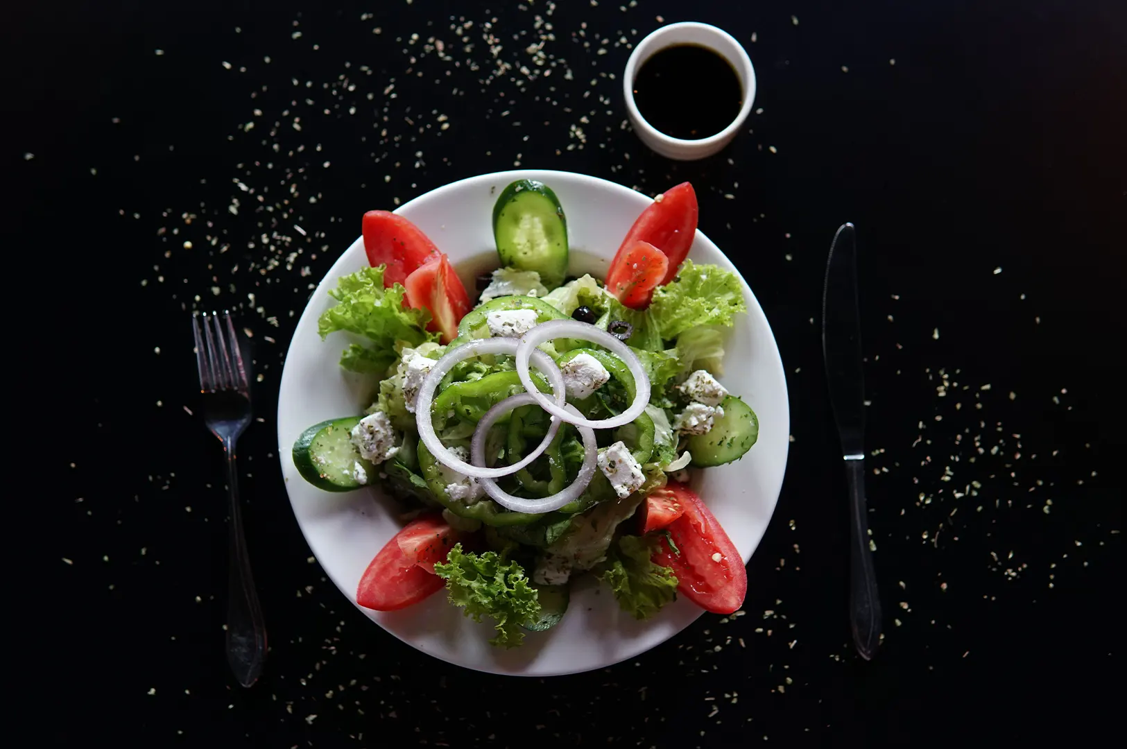 Fresh Greek salad with tomatoes, cucumbers, feta cheese, and onions on a white plate with a side of dressing on a black table.