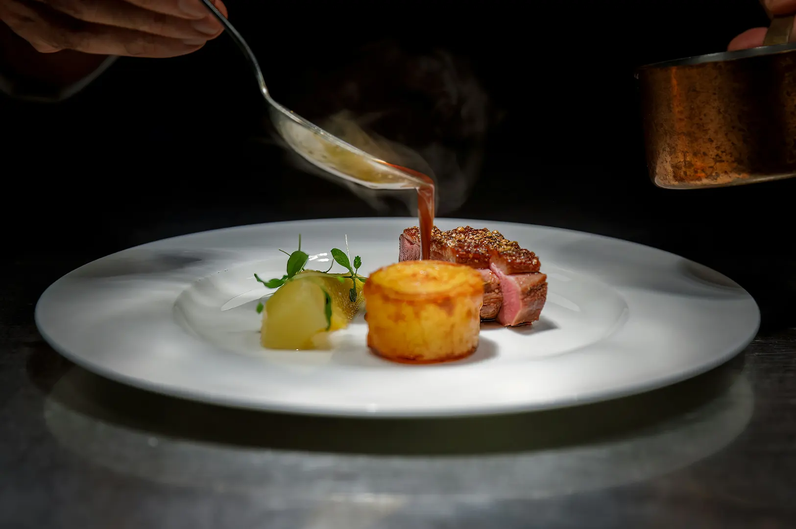 Chef pouring sauce over gourmet duck breast and potato gratin on a white plate.