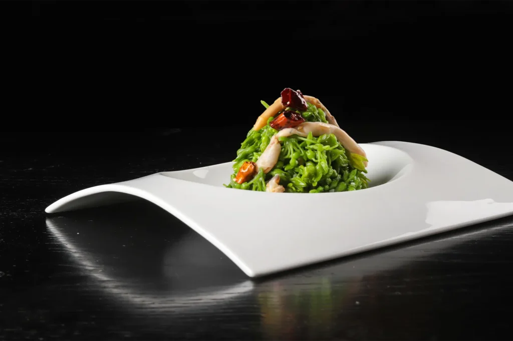Gourmet green vegetable salad with glossy, oil-touched texture served on a modern white plate, captured with dramatic studio lighting against a black background.