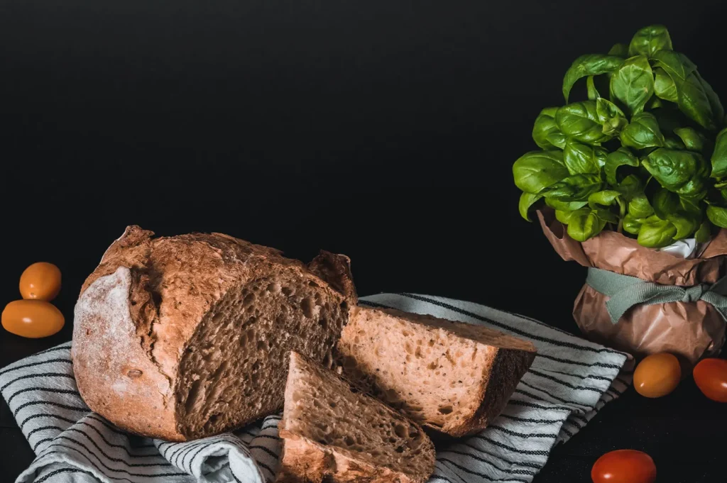 Dark moody food photography of freshly baked artisan bread sliced on a striped kitchen towel, accompanied by fresh basil and cherry tomatoes against a black background.