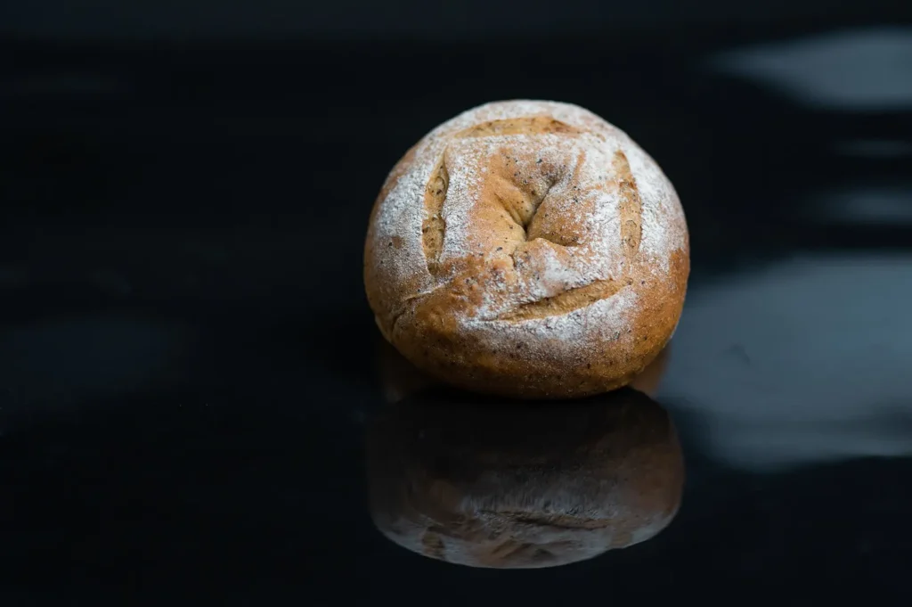 Freshly baked artisanal round bread roll dusted with flour on a reflective dark surface.