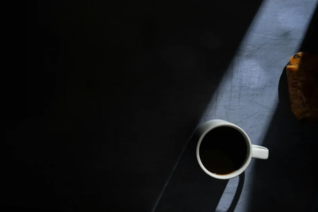 A white mug of black coffee and a pastry on a table in dramatic side lighting.