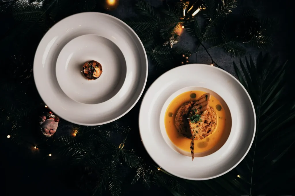 Elegant holiday dish setting with two white plates centered; left plate has a small appetizer, right plate features a main dish in creamy sauce. Surrounded by pine branches, ornaments, and warm string lights for a festive ambiance.