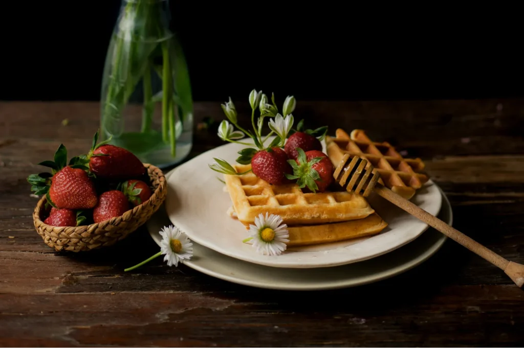 A rustic setting with golden waffles topped with fresh strawberries and white flowers on a plate. A honey dipper rests nearby; a vase with leaves and a basket of strawberries are in the background.