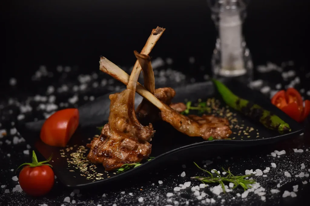 Grilled lamb chops served on a black plate with fresh tomatoes and herbs against a dark background.
