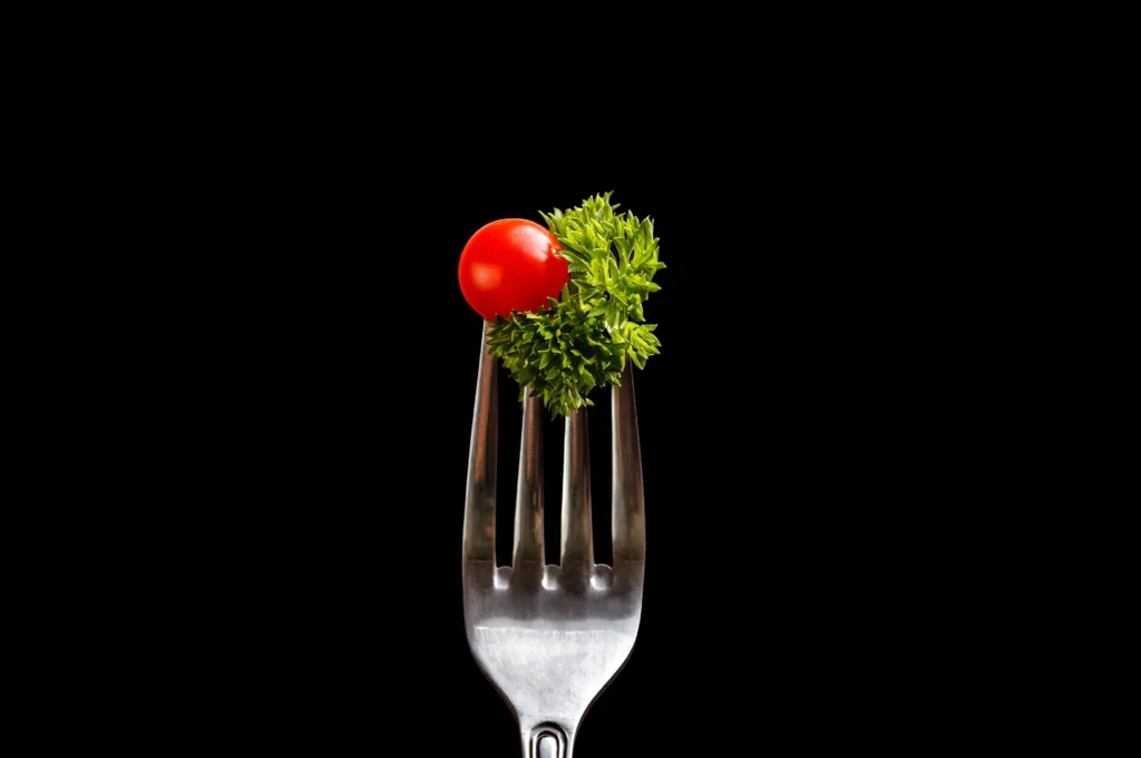 Close-up macro food photography of a fresh cherry tomato and crisp green parsley on a silver fork against a dark black background.