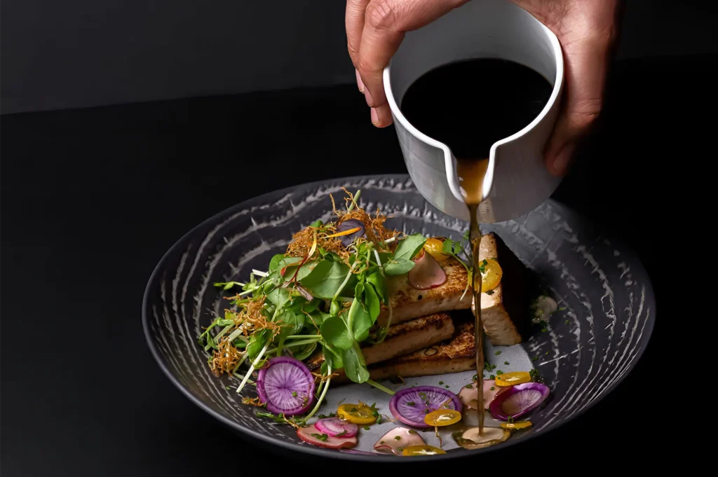 Professional food photography of sauce being poured over a gourmet tofu and microgreens dish in a dark bowl.