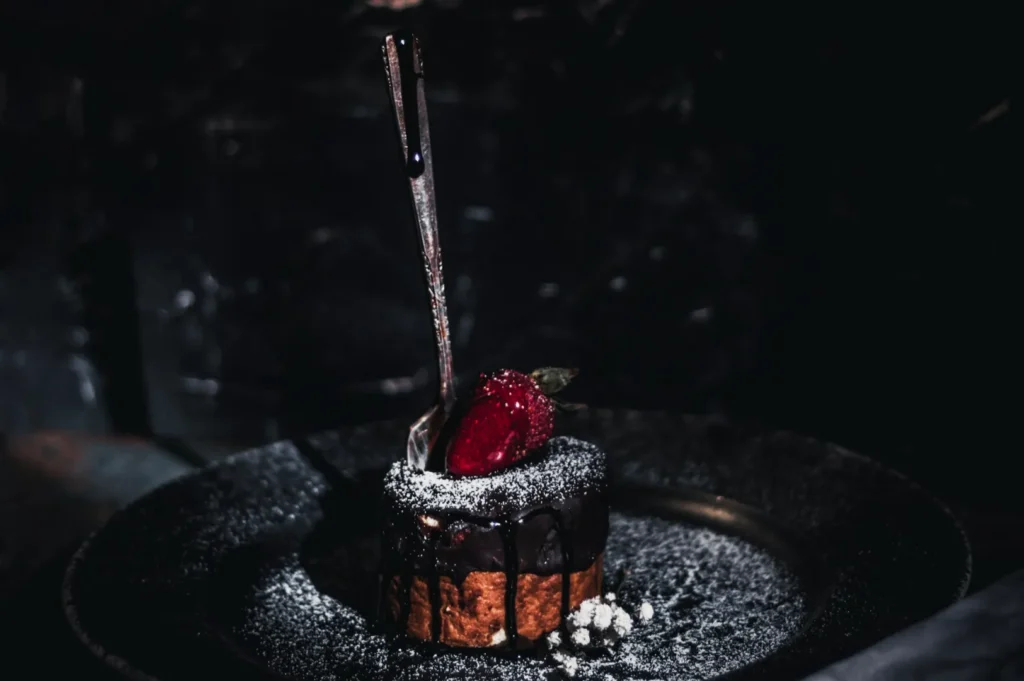 A chocolate cake on a dark plate, topped with a strawberry and drizzled chocolate, dusted with powdered sugar. A fork is elegantly standing upright, creating a moody, indulgent atmosphere.