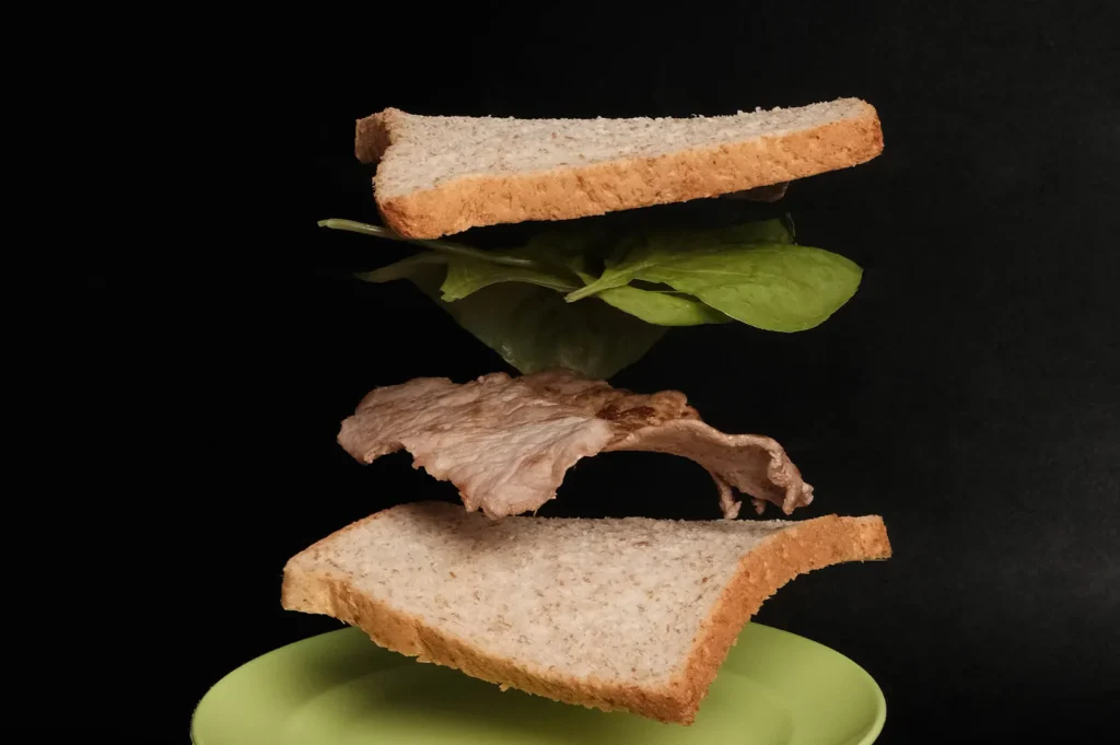 Floating sandwich ingredients including whole wheat bread, fresh spinach, and sliced meat above a green plate against a black background.
