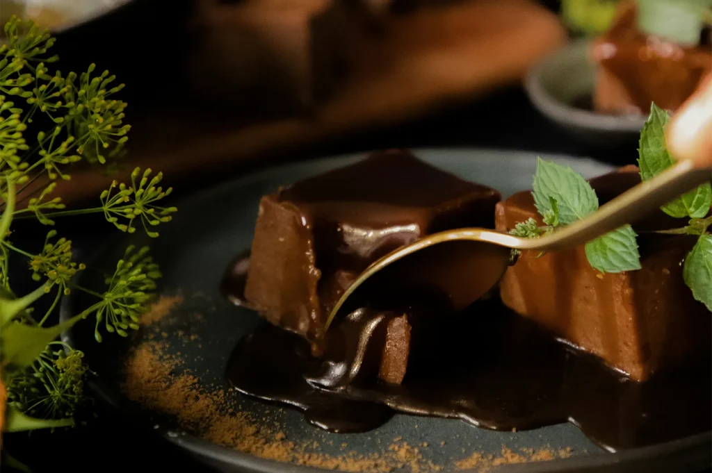 A close-up of a decadent chocolate square dessert with a shiny, reflective chocolate sauce glaze, garnished with fresh mint and being lifted by a gold spoon.